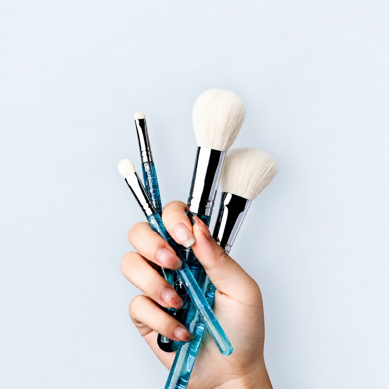 A hand holding four actual ice-blue brushes, showing different head shapes and clear acrylic handles with silver floral decoration
